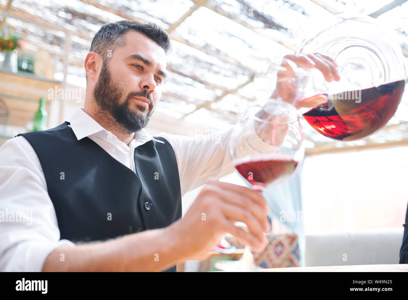 Young bearded barman or sommelier pouring red wine into wineglass Stock Photo