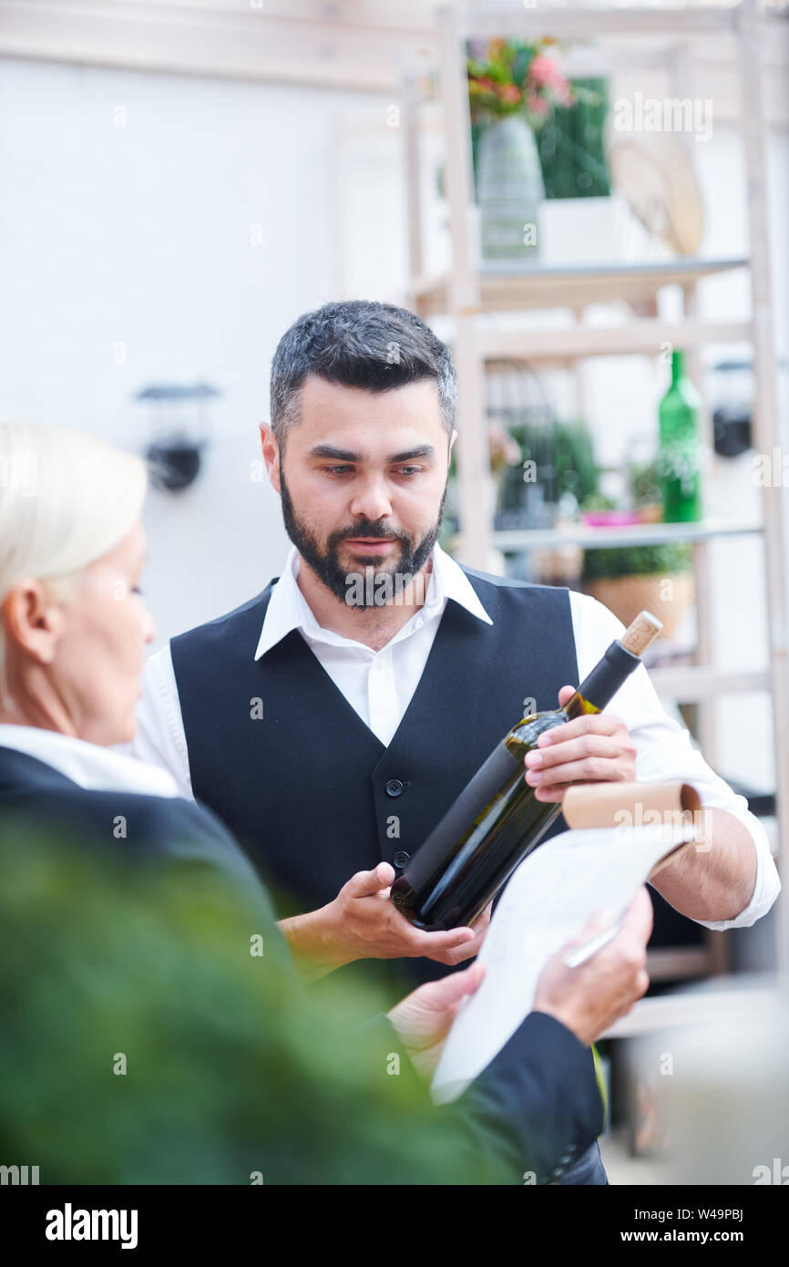 Young bartender giving characteristics of new sort of wine in bottle to cavist Stock Photo