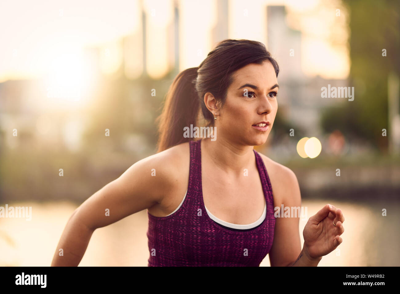 Portrait of active millenial woman jogging at dusk with an urban cityscape and sunset in the background Stock Photo