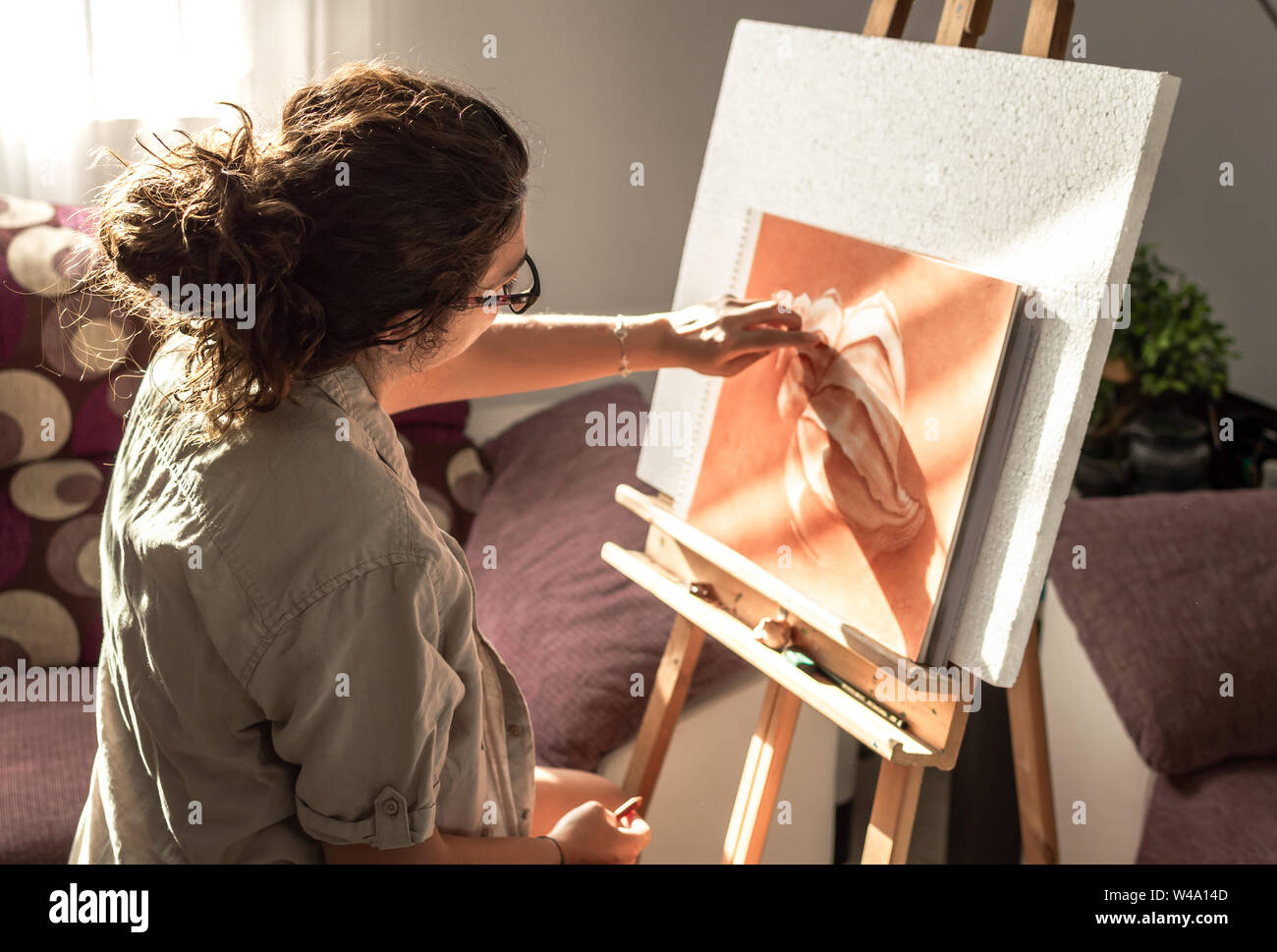 Woman with casual clothes and glasses painting with sanguine or red chalk. Drawing at home in front of a sunny window with easel. Stock Photo