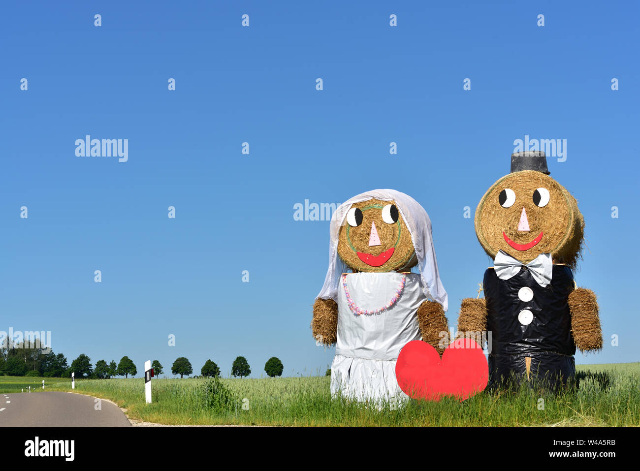 two traditional huge straw dolls in the form of a wedding couple stand in Bavaria on the roadside against a blue sky Stock Photo