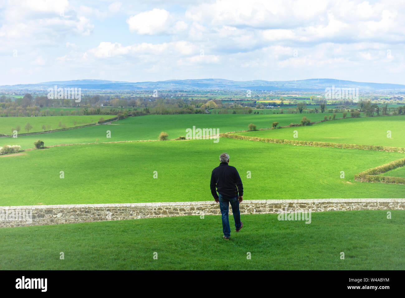 Middle-aged man with dark clothes, walking across a field, before to jump a wall, a stone fence. Overcome difficulties. Walk facing towards. Looking a Stock Photo