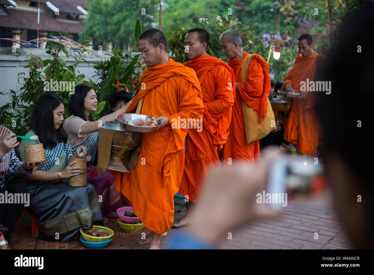 Tourist taking a photo of people giving alms to Buddhist monks on the street early in the morning in Luang Prabang, Laos. The ritual is called Tak Bat Stock Photo