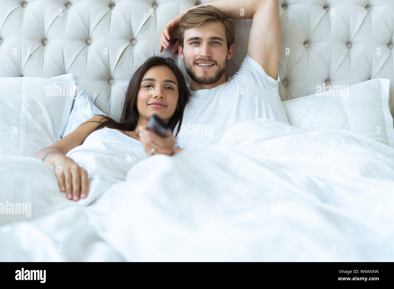 happy couple with remote lying in bed at home and watching tv Stock