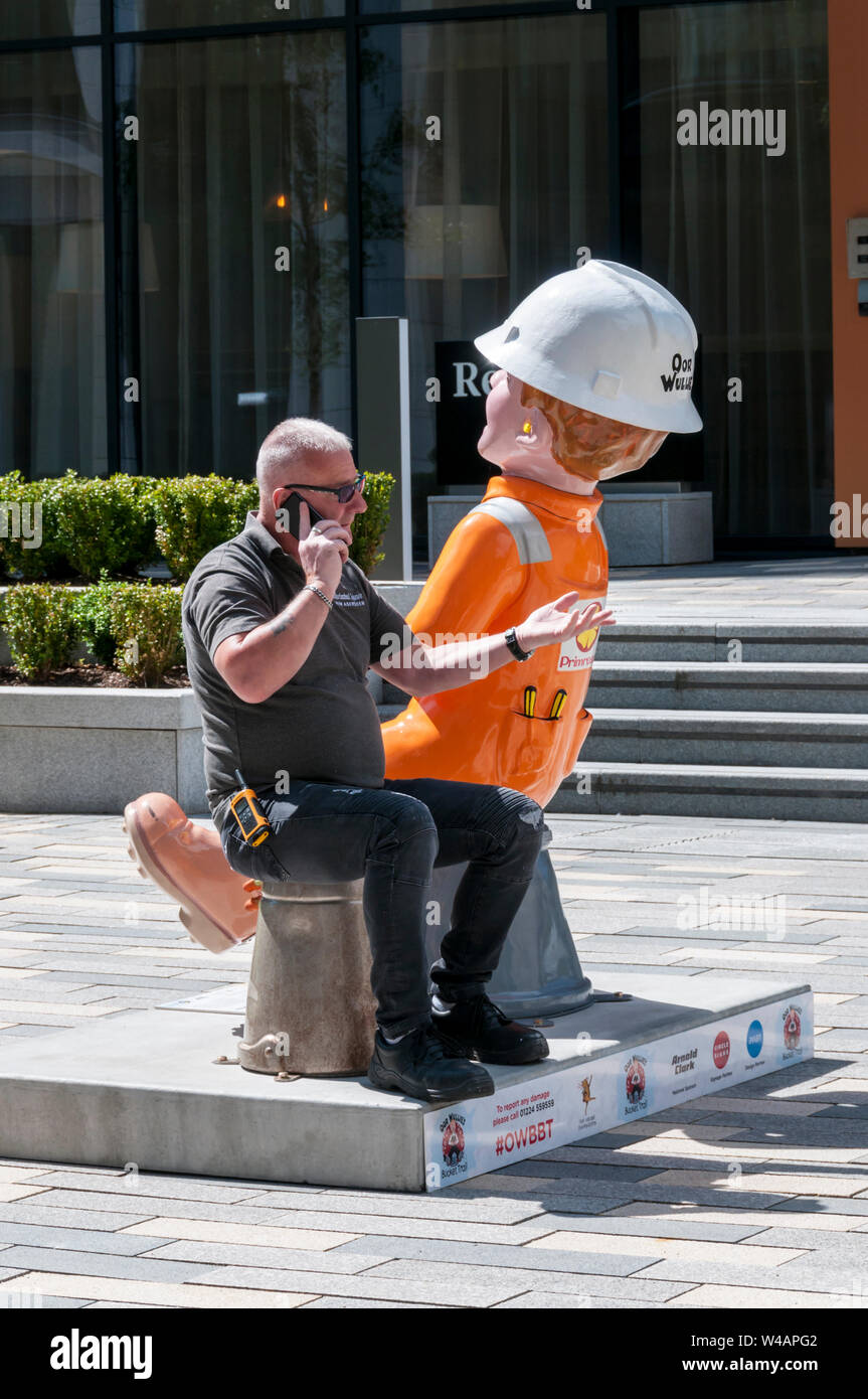 Life imitates art as a man sits on the upturned bucket of an Oor Wullie statue in Aberdeen. Stock Photo