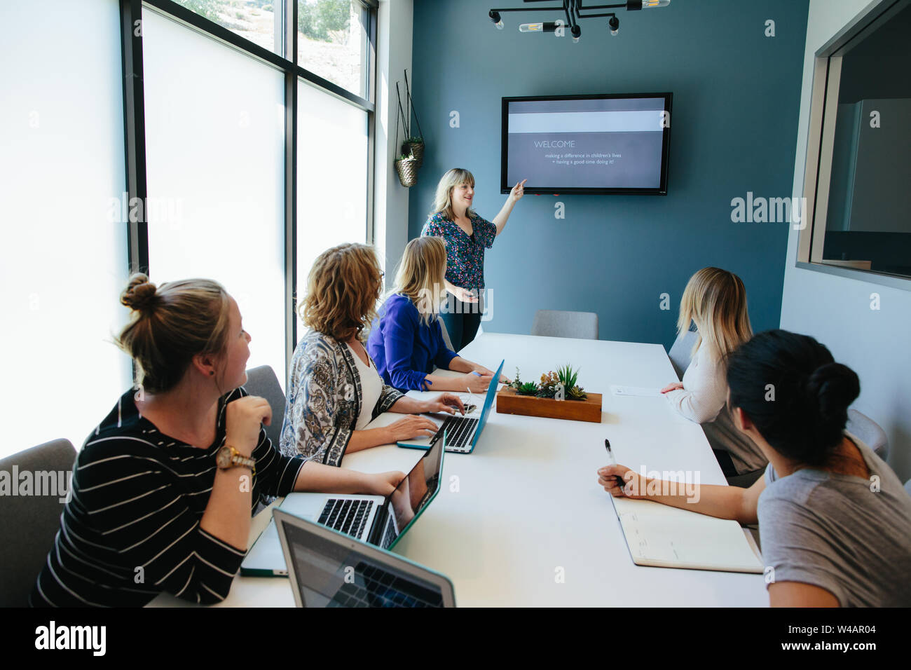 Women sitting at a conference table look and listen to woman standing Stock Photo