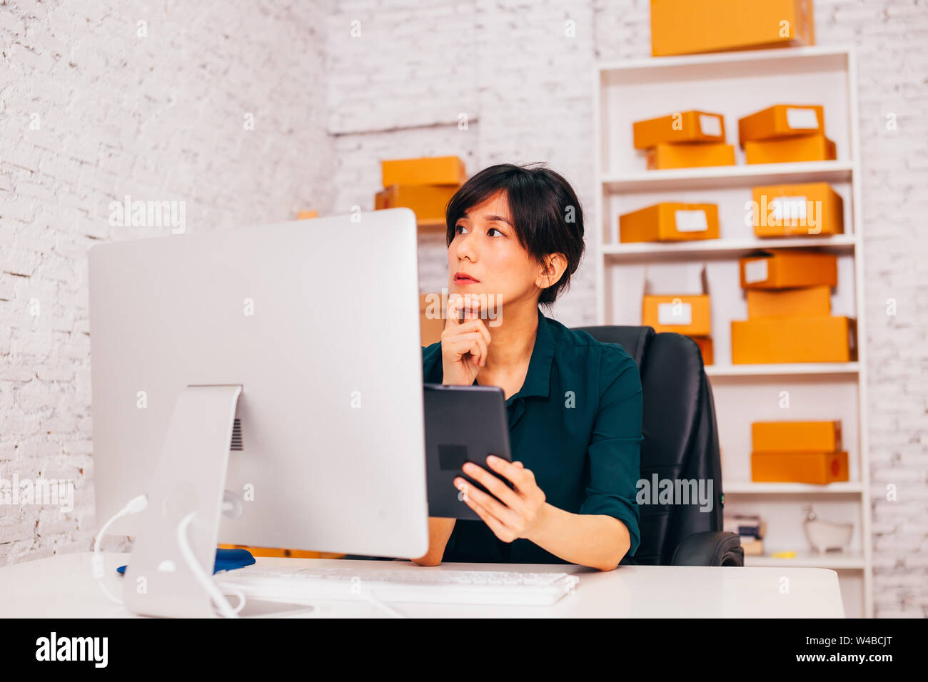 Asian entrepreneur with tablet looking away and thinking while sitting at desk in office of e commerce company Stock Photo