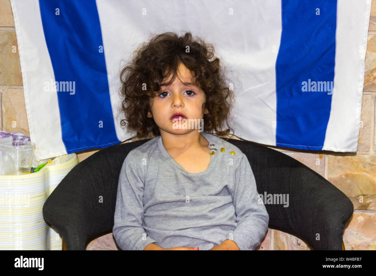 5 May 2018 A pretty young Jewish girl sits quietly in front of an Israeli Flag as her parents attend to customers in their restaurant in Nazareth Stock Photo