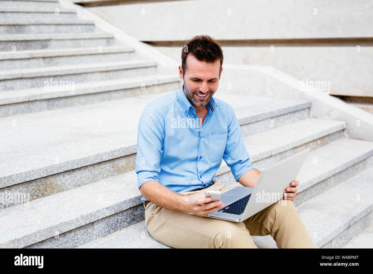 Handsome student sitting on stone steps and using a laptop and mobile Stock Photo