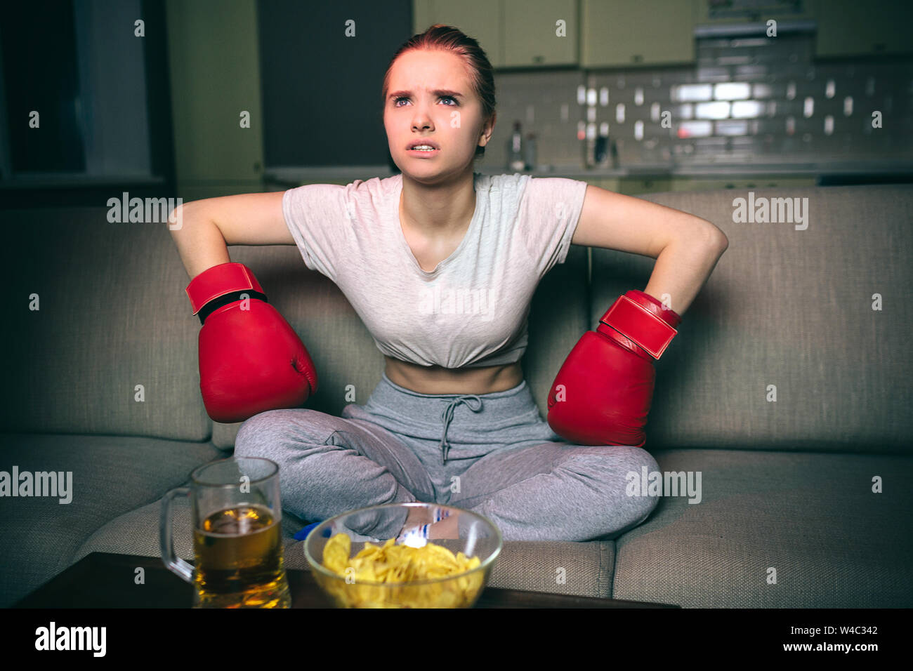 Young woman watch boxing on tv at night. Serious upset model sit on sofa and lean forward. Hands in sport gloves