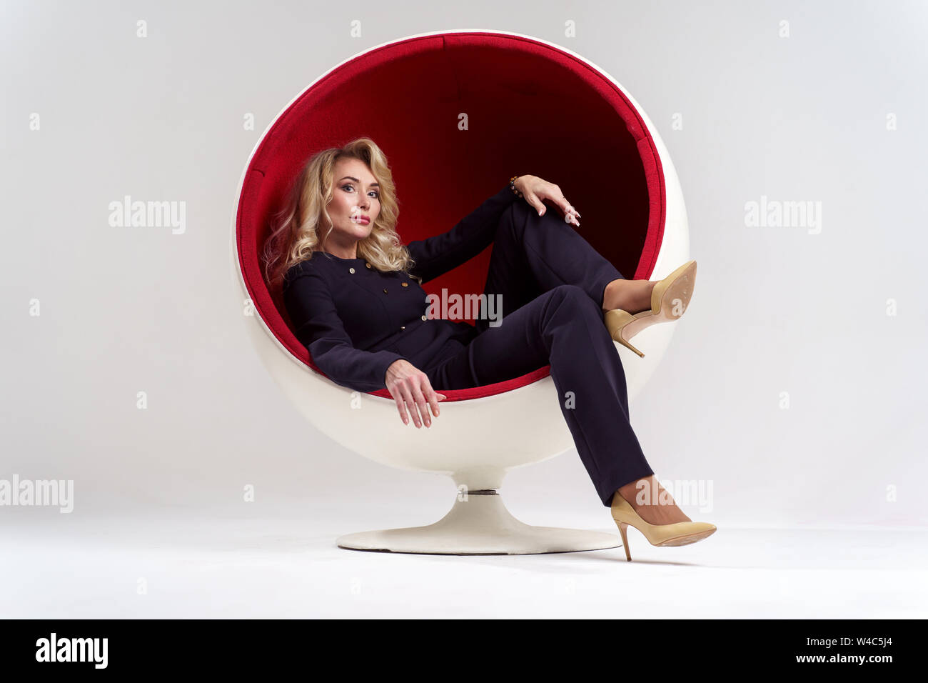 Photo of blonde woman with long curly hair in black suit sitting in round chair isolated on white background Stock Photo