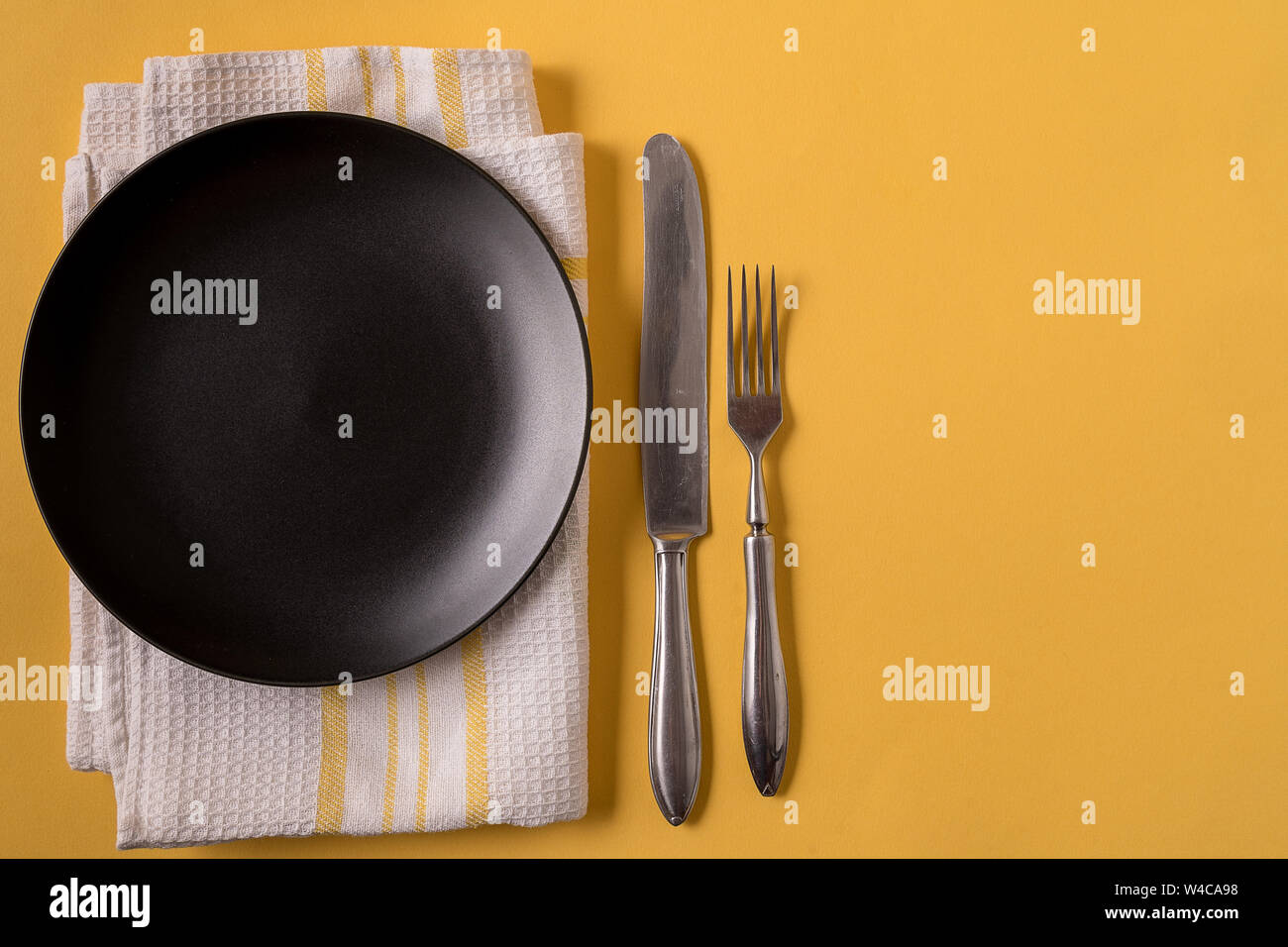 Flat lay food background with empty black plate, cutlery and napkin, over yellow background, copy space. Stock Photo