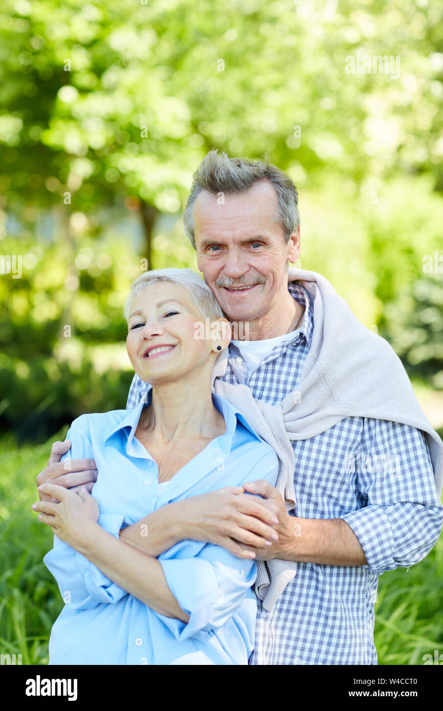 Waist up portrait of happy senior couple embracing and looking at camera while enjoying walk in Summer park Stock Photo
