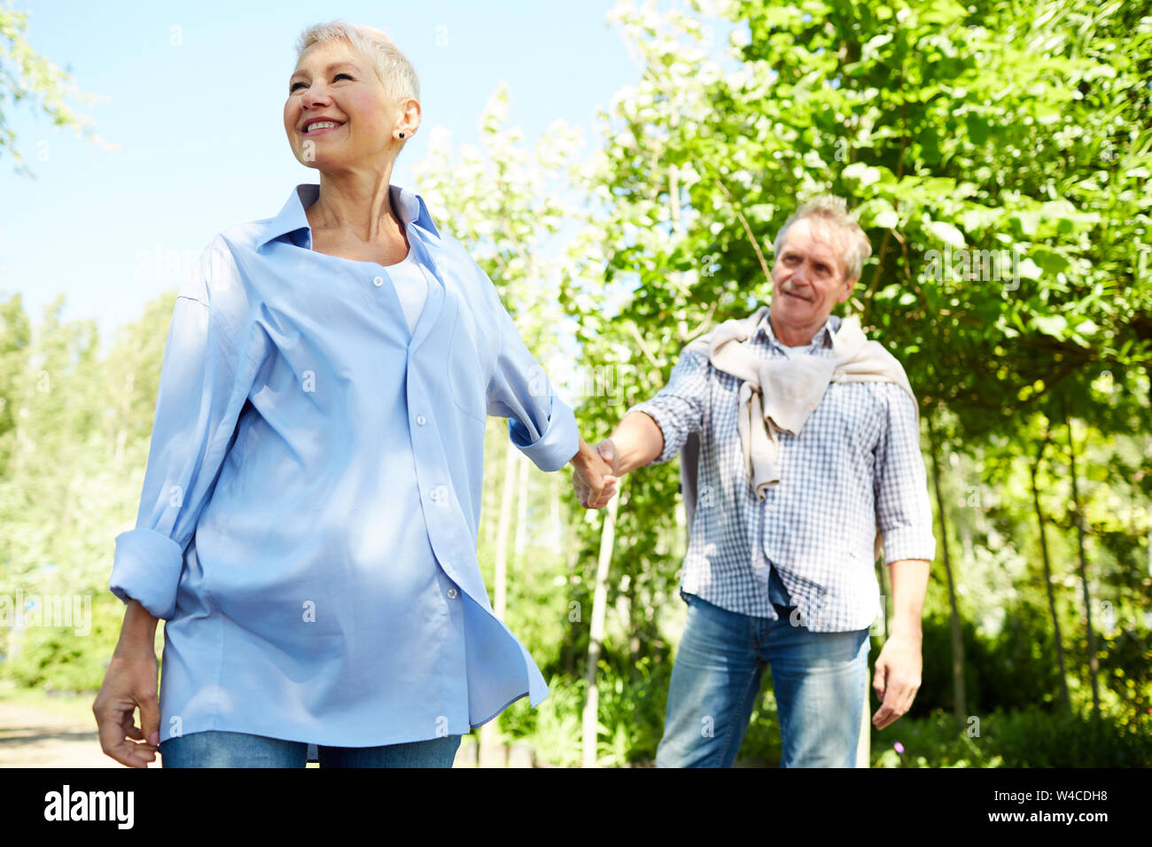 Portrait of happy senior woman leading husband by hand while enjoying walk in Summer park Stock Photo