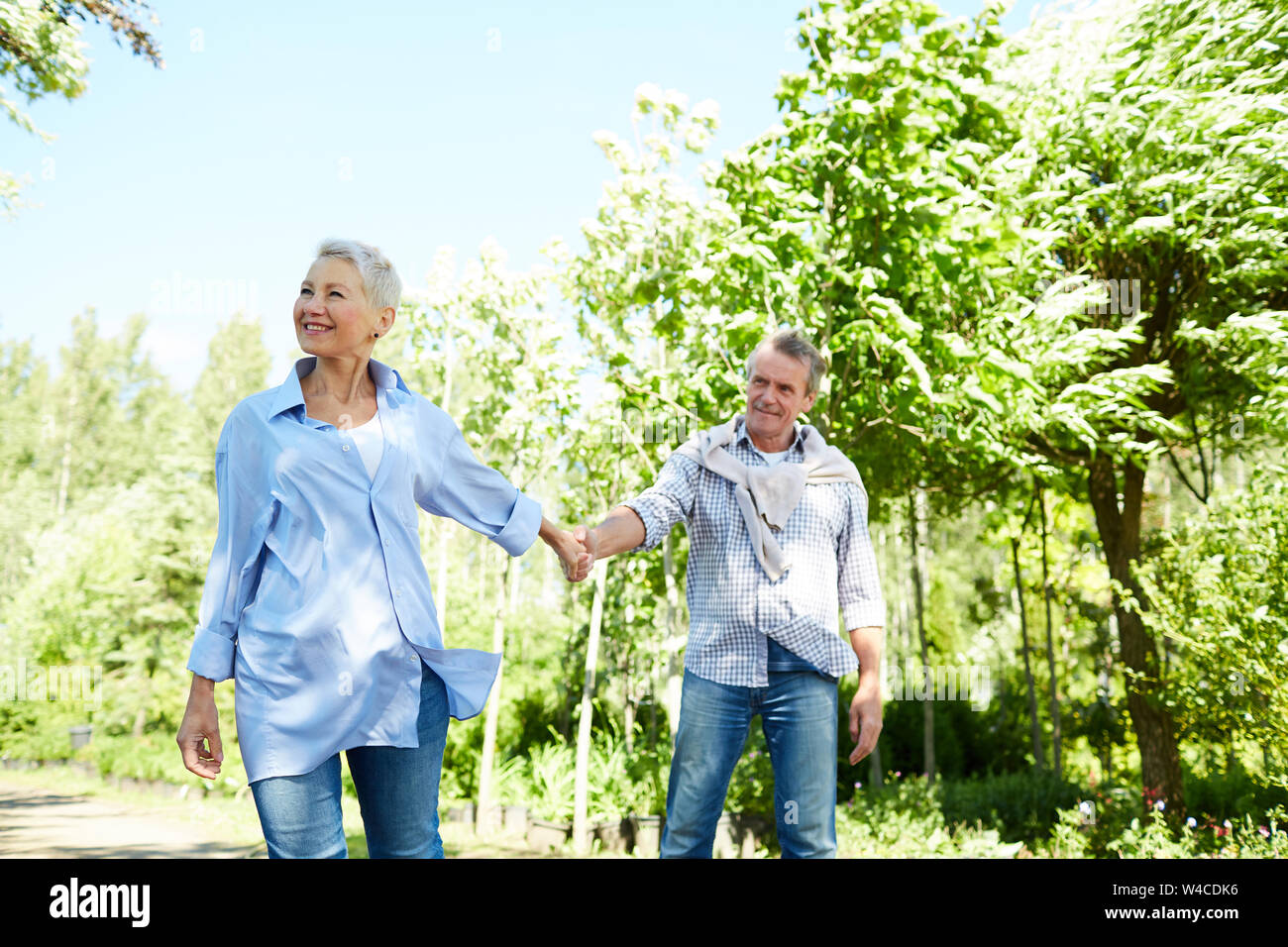 Portrait of happy senior woman leading husband by hand while enjoying walk in Summer park, copy space Stock Photo