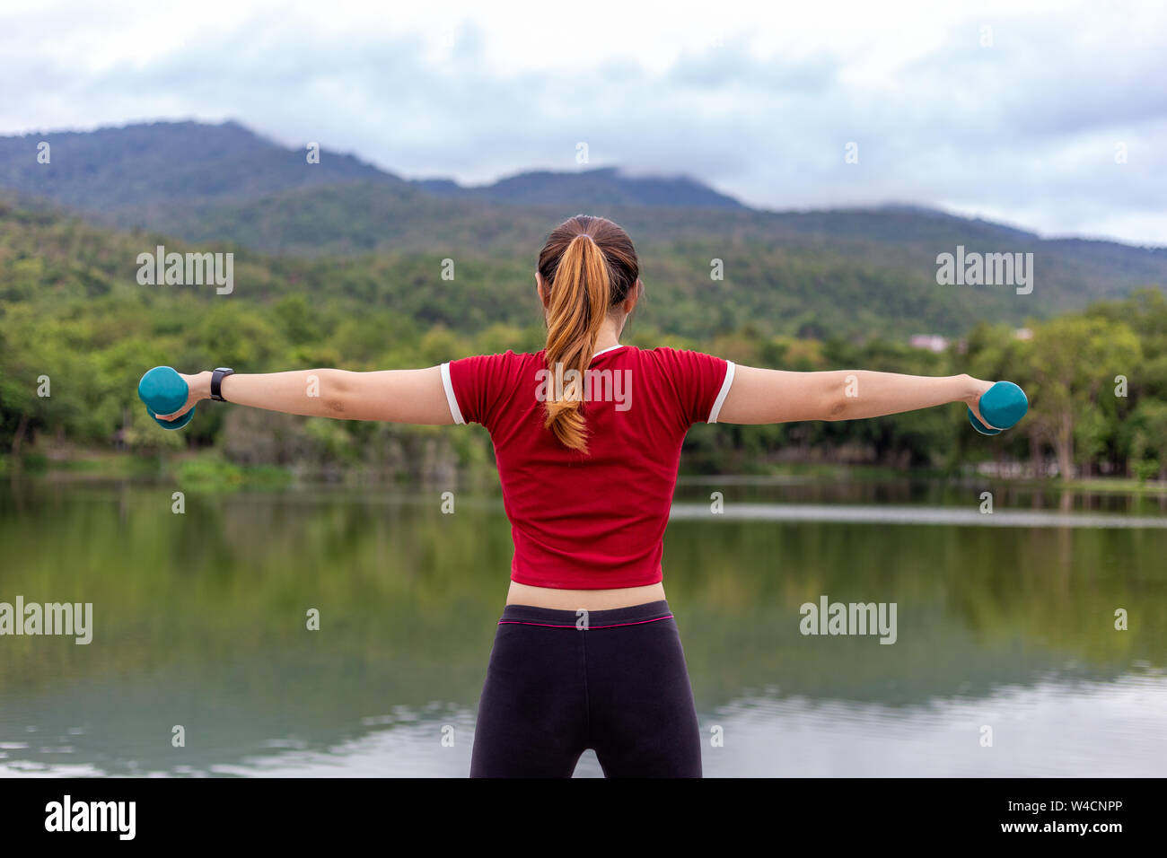 Back view of young athlete woman lifting dumb bells with both arms during her morning  exercise at a lake park with view of lake and mountains in the Stock Photo