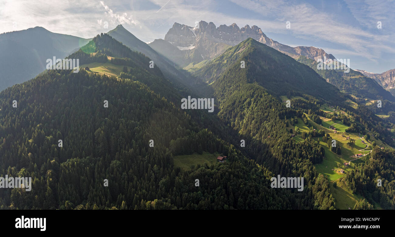 aerial images over an alpine valley and lush green pine forests with chalets and high mountains surrounding the valley. Stock Photo