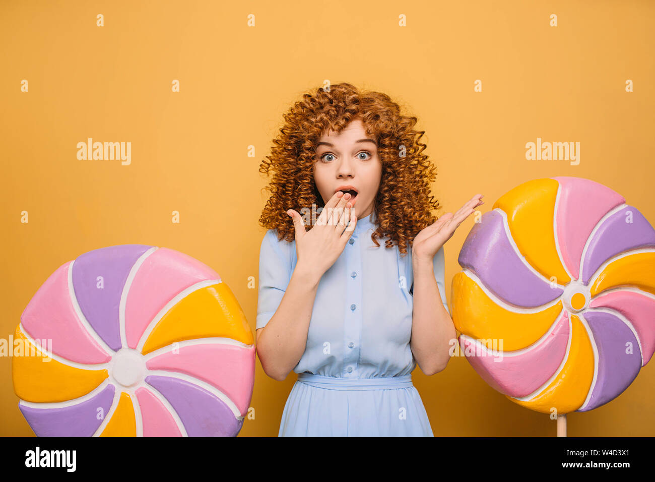 Funny woman with curls and emotionally shocked face standing near big lollipops Stock Photo