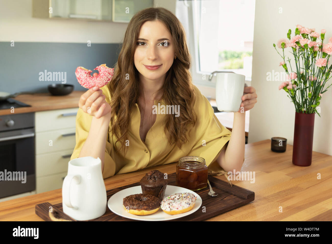 Young beautiful girl is having breakfast at home in the kitchen. She drinks her morning coffee and with great pleasure eats a donut with pink icing. Stock Photo