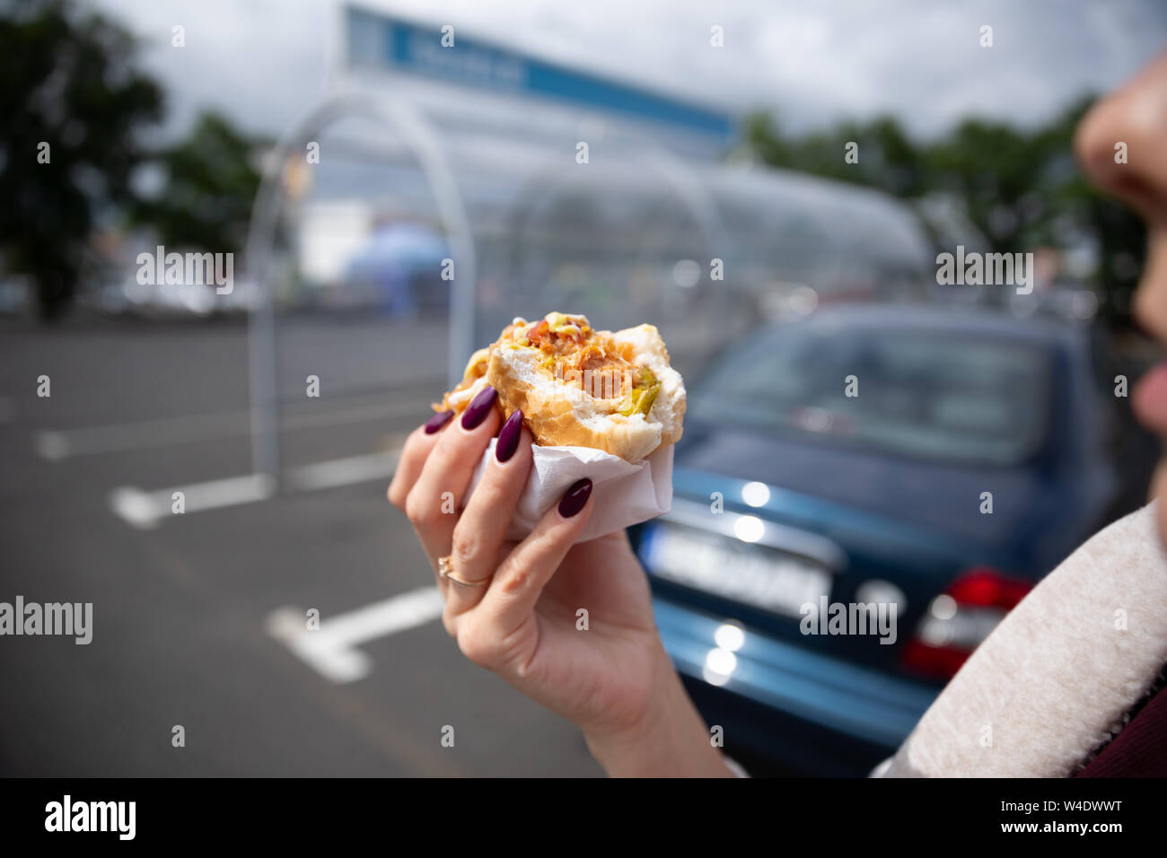 A young woman holds a bitten hot dog in her hand. Snack in the parking lot near the shopping center. Stock Photo