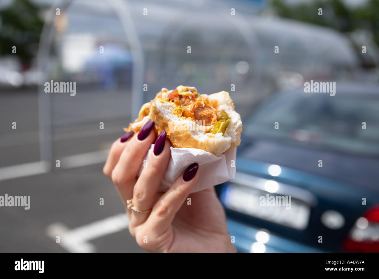A young woman holds a bitten hot dog in her hand. Snack in the parking lot near the shopping center. Stock Photo