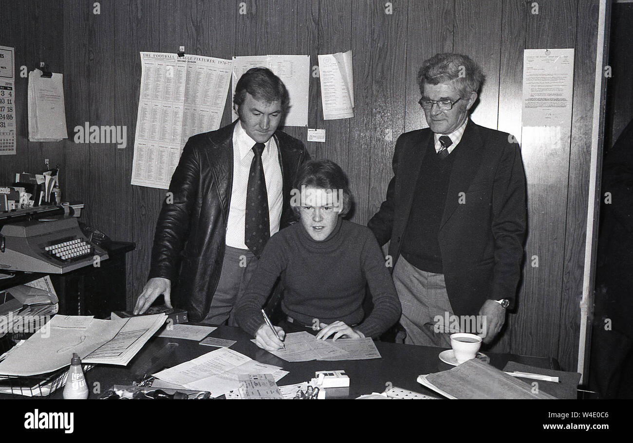 1976,  historical, a young football player signing his contract with Millwall Football Club in the club secretary's office. Of note are the clothing fashions of the day, and the typewriter, paperwork and a packet of cigarettes on the desk. Stock Photo
