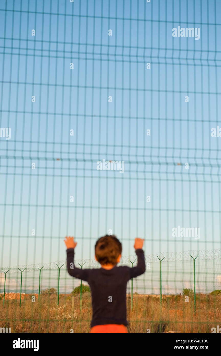 one helpless little child refugee standing near border holding fence with razor wire Stock Photo