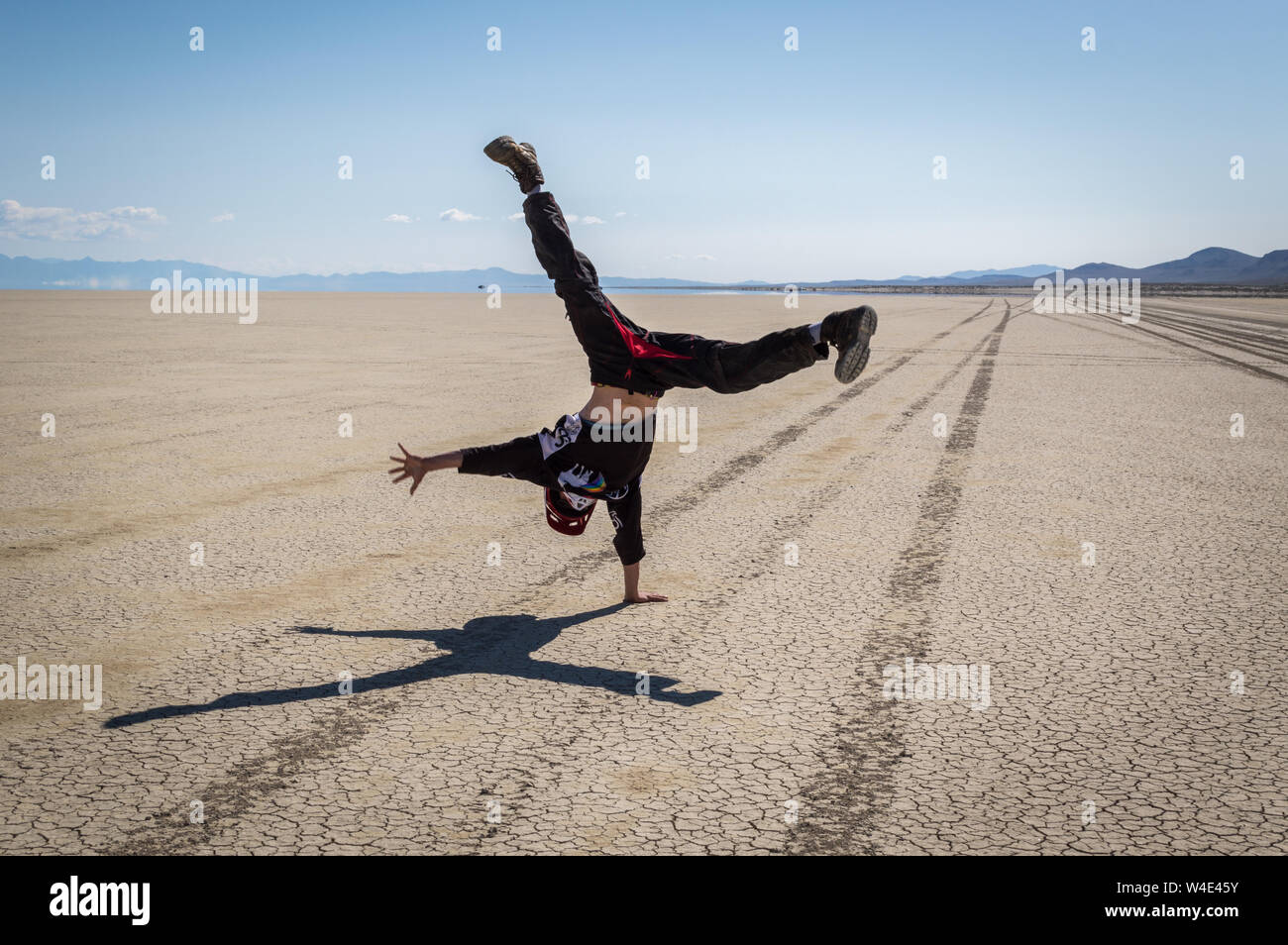 A young man does a cartwheel on the Black Rock Desert with tire tracks leading in the distance on a sunny day. Stock Photo