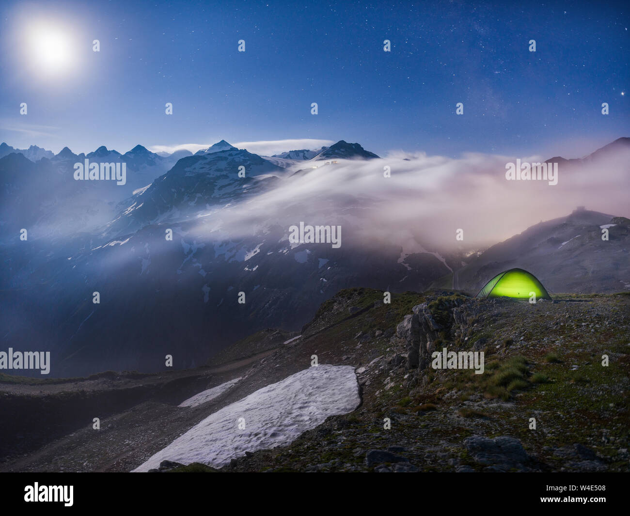 Moonlit scene with illuminated green tent under starry night - fog rolling over mountains Stock Photo