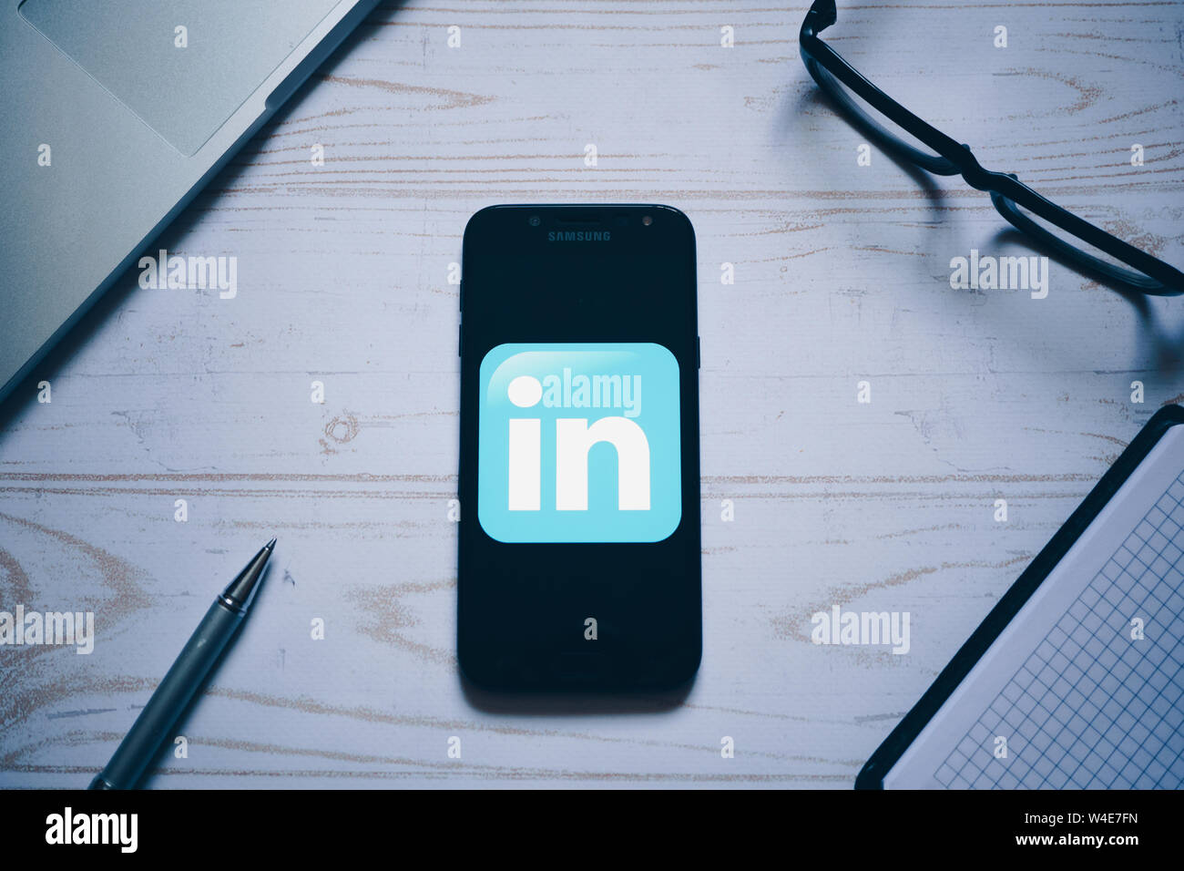 Nizhyn, Ukraine/ July-16-2019: LinkedIn app logo on the screen of modern smartphone. Flat lay. Laptop, glasses, notebook and pen on background. Social Stock Photo