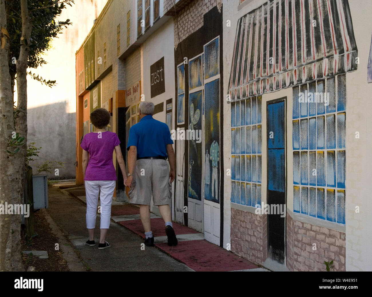 Caucasian Senior Couple (age 60-70) walking down sidewalk with painted mural on wall at  Georgetown SC, USA. Stock Photo