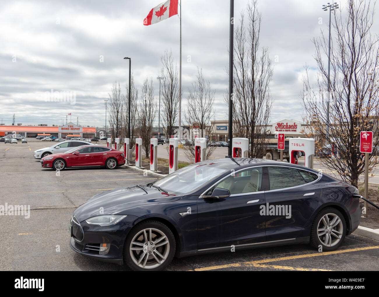 Blue Tesla Model S parked, supercharging in Markham, ON with Model S, Model X and Canadian Flag waving in the background. Stock Photo