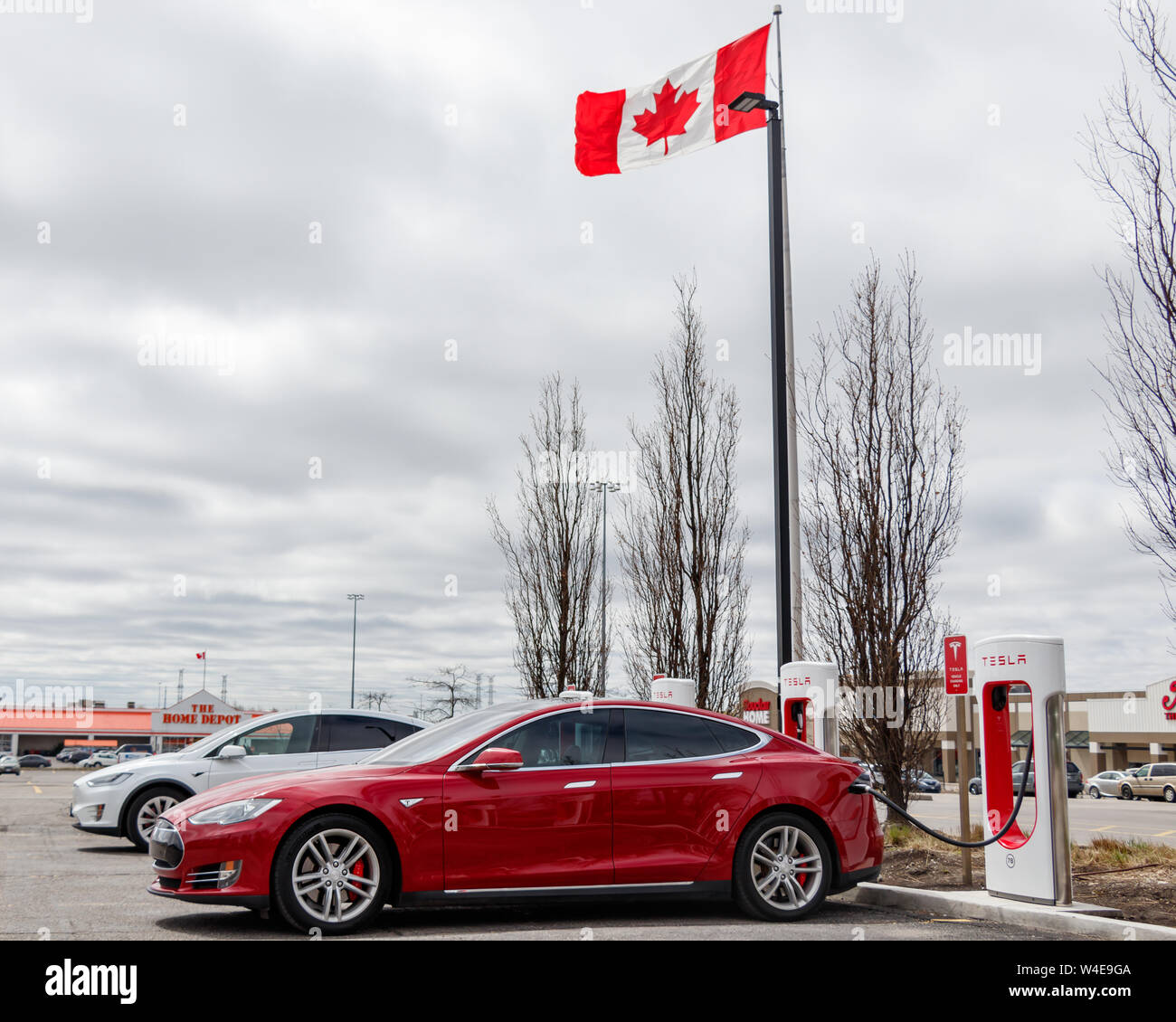 Red Tesla Model S parked, supercharging at SmartCentres Markham Woodside with white Model X and Canadian Flag waving. Stock Photo