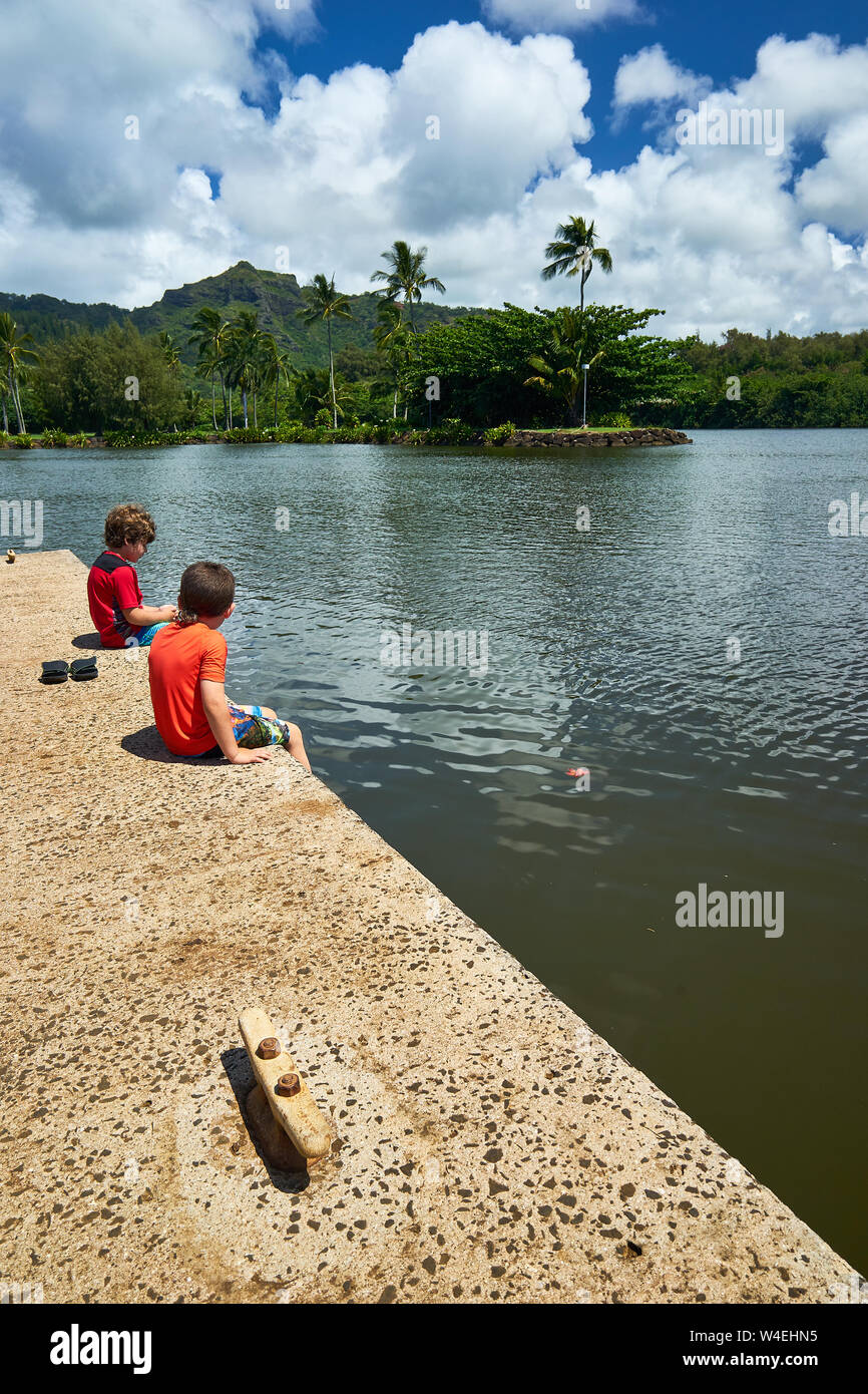 Two brothers sit together on a concrete platform at Wailua River with palm trees and green mountain in the background at Kauai, Hawaii. Stock Photo