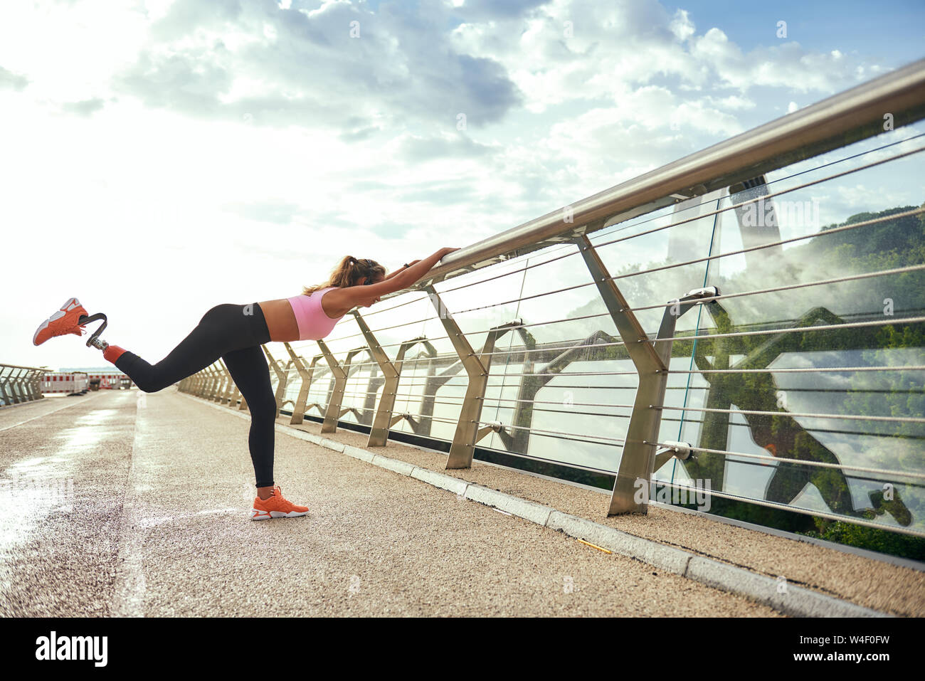 Morning workout. Disabled athletic woman with prosthetic leg in sportswear doing sport exercises while standing on the bridge. Side view. Disabled sport concept. Motivation Stock Photo