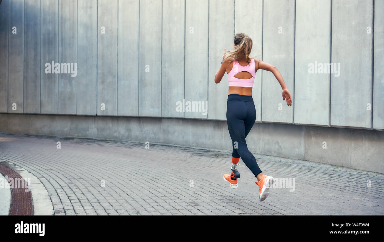 On the way to success.Back view of young disabled woman with leg prosthesis in comfortable sports clothing is running outdoors along the street. Sport concept. Motivation. Healthy lifestyle Stock Photo
