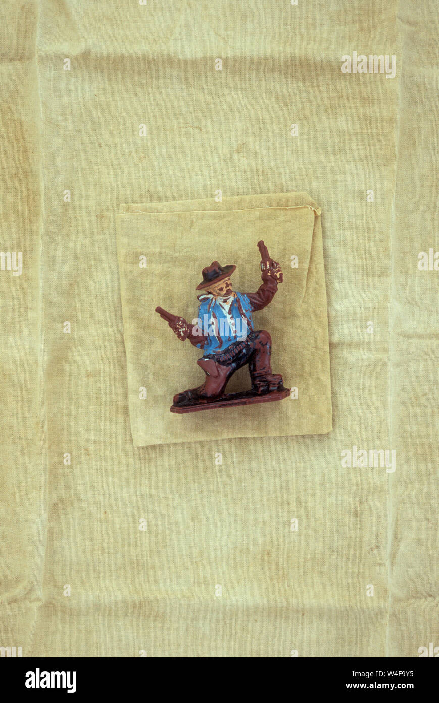 Plastic model of cowboy in blue jacket kneeling and waving or shooting guns with both hands lying on stained and folded linen Stock Photo
