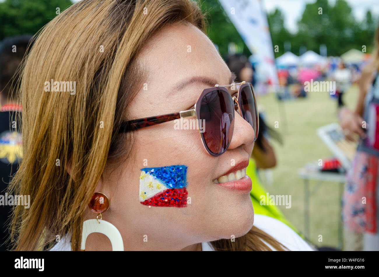 A Filipino lady wearing sunglasses with the flag of The Philippines painted with face paint and glitter on her cheek. Stock Photo