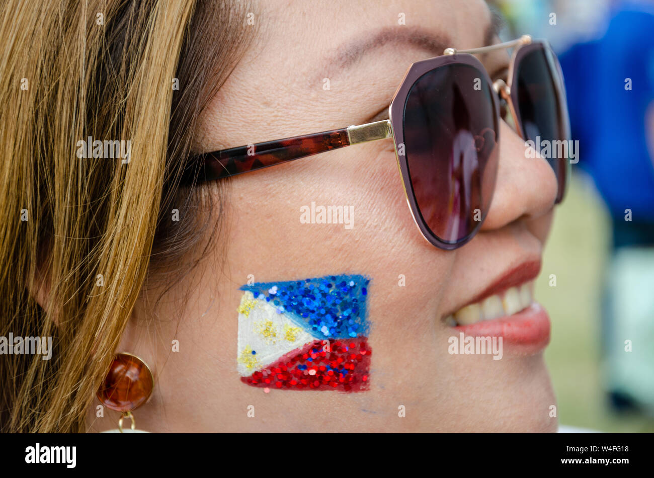A Filipino lady wearing sunglasses with the flag of The Philippines painted with face paint and glitter on her cheek. Stock Photo