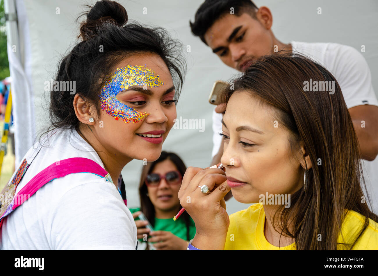 A lady sits and has face paint applied. The artist applying the face paint has face glitter around her eye. Stock Photo