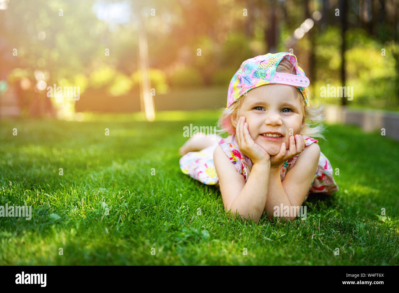 little girl laying on green grass with hands on cheeks at home backyard on sunny summer day Stock Photo