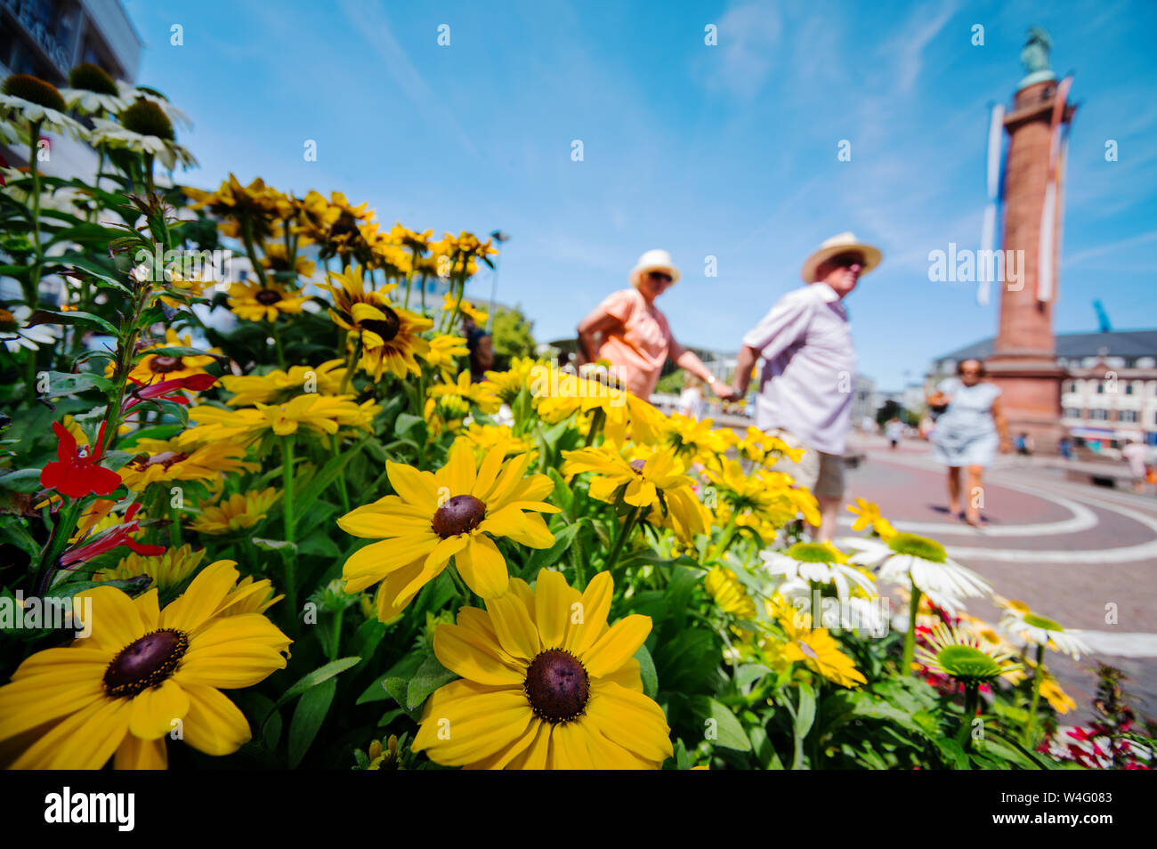 Darmstadt, Germany. 23rd July, 2019. Two people in a sun hat walk past a flower stand. In the background is the Darmstadt landmark of the 'Lange Ludwig'. Credit: Andreas Arnold/dpa/Alamy Live News Stock Photo