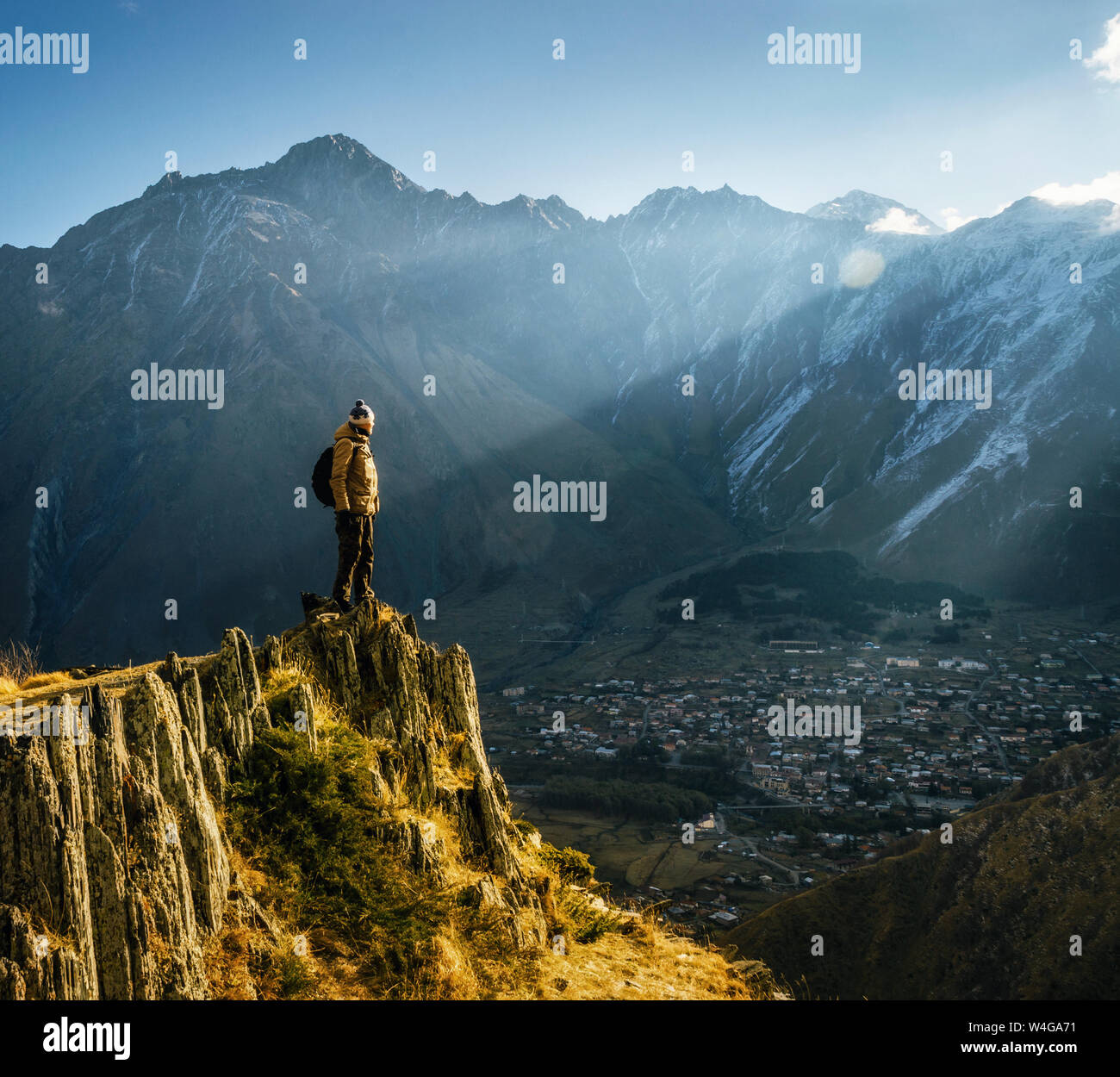 Young tourist in bright hat, black trousers with a backpack stands on cliff's edge and looking at the misty mountain village and glacier at sunrise Stock Photo