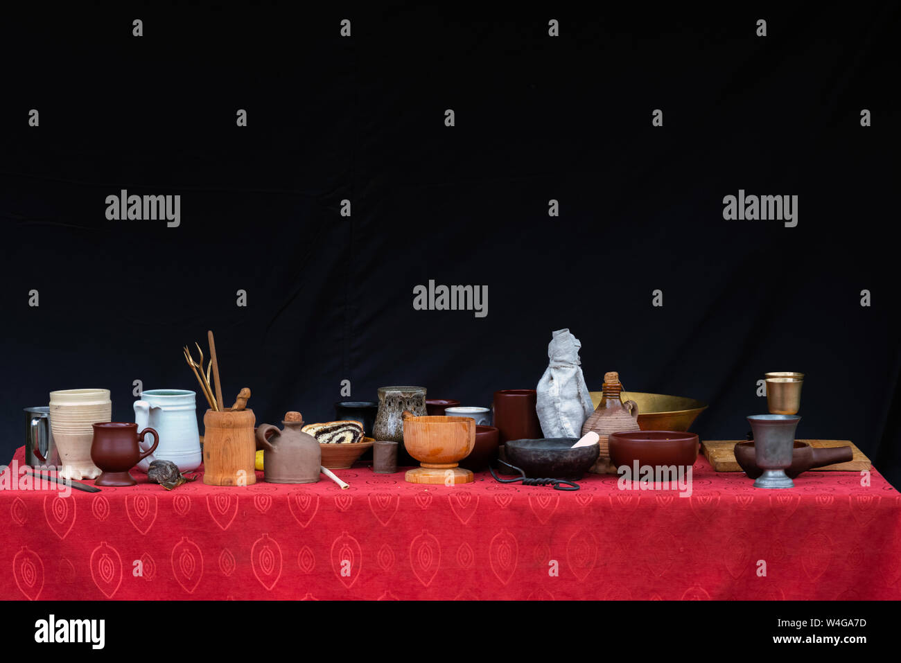 A lot of clay cups, pots, plates, vases and other dishes on top of table with red tablecloth and black background Stock Photo