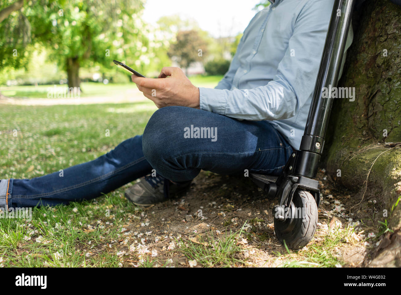 Man sitting on E-Scooter in a park leaning against tree trunk while using smartphone, partial view Stock Photo