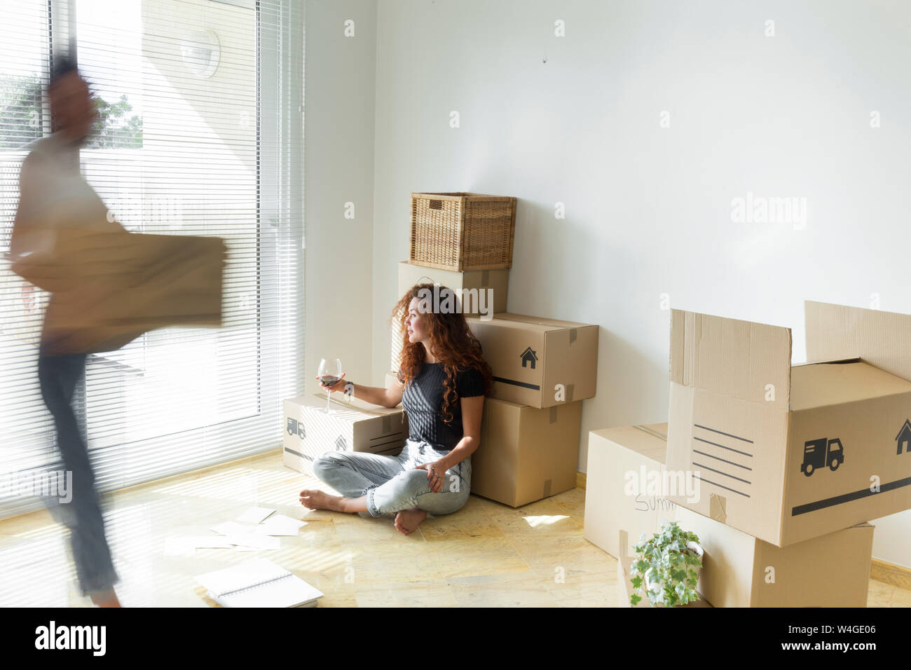 Young woman sitting in new home with glass of red wine while friend is moving fast with cardboard box Stock Photo