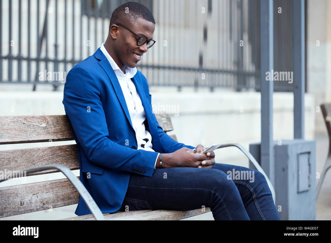 Young businessman wearing blue suit jacket sitting on bench and using smartphone Stock Photo