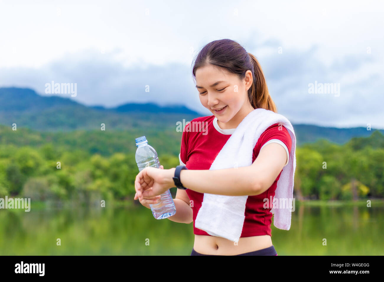 Young happy athlete woman holding a bottle of water and looking at her smart watch to check her exercise progress after her morning routine at a lake Stock Photo