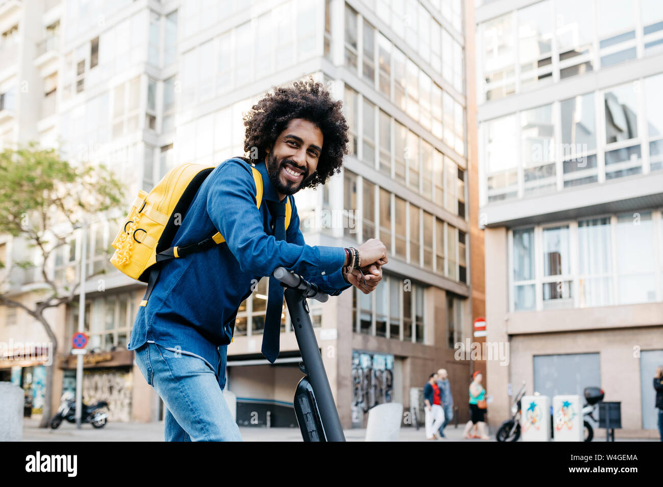 Portrait of smiling young man with yellow backpack on E-Scooter in the city, Barcelona, Spain Stock Photo