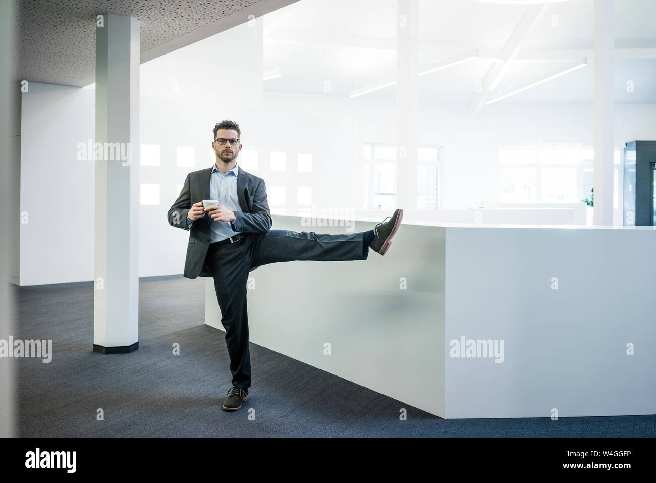 Businessman with cup of coffee standing on one leg in office Stock Photo